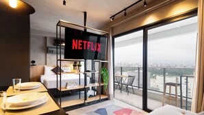 A modern hotel room with a large window, a balcony, a dining area with plates and glasses, and a TV screen displaying "NETFLIX". - Nun Vila Nova 2503 - Vila Nova Conceição (São Paulo)
