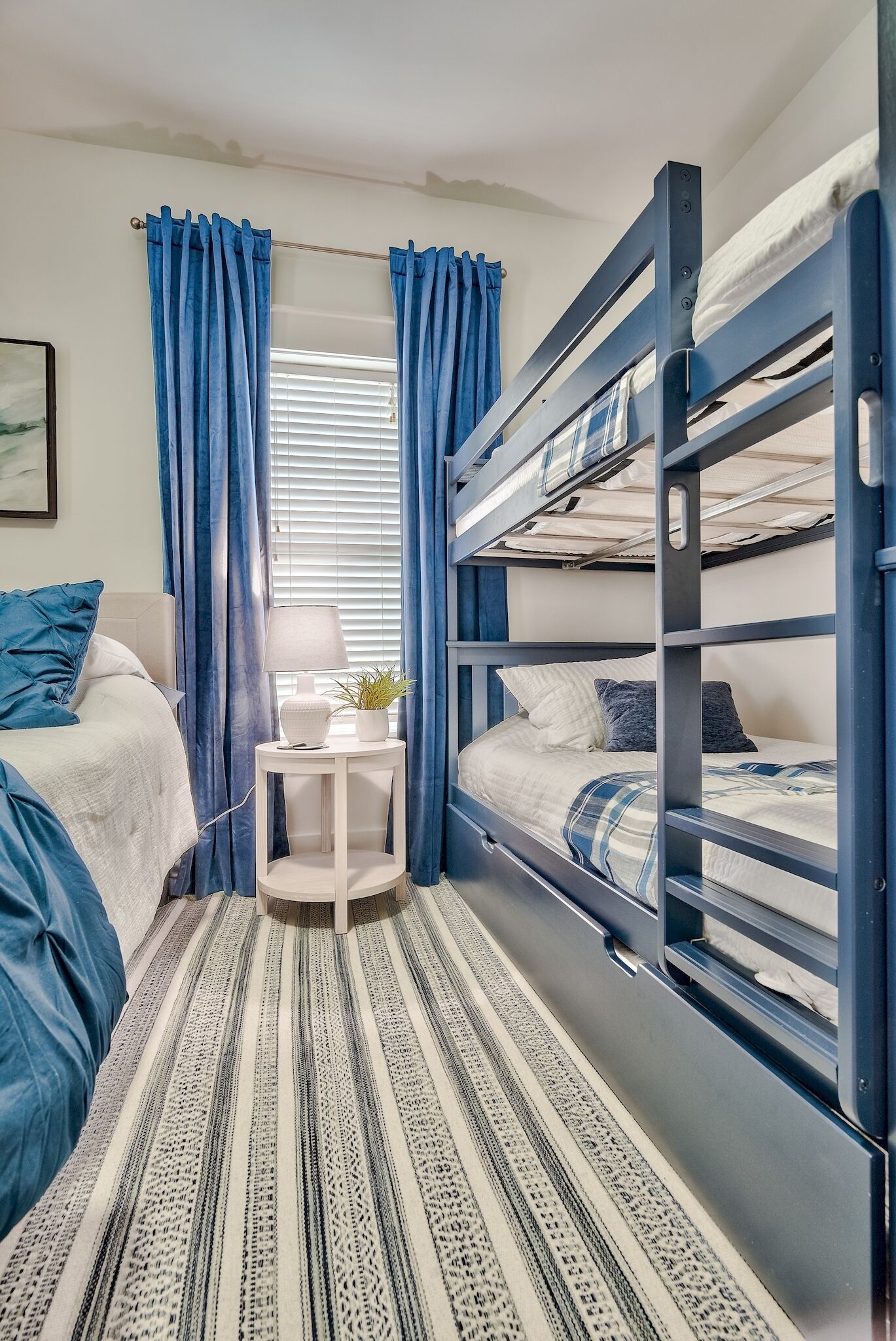 A bunk bed room with a striped carpet, a bedside table with a lamp and plant, and blue curtains.
