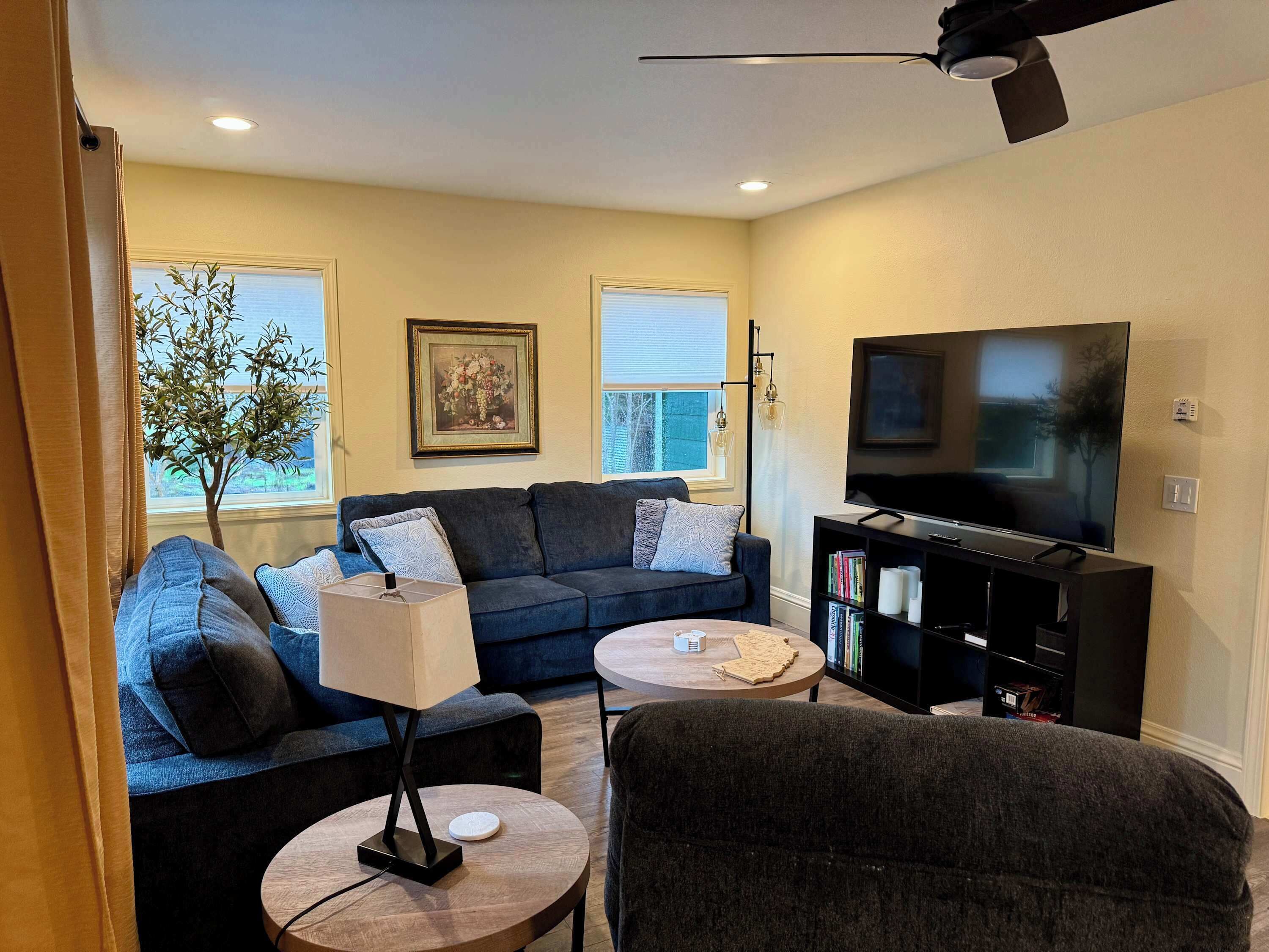 A modern living room with a sofa, coffee table, lamp, TV, bookshelf, and a large window with a view of trees.