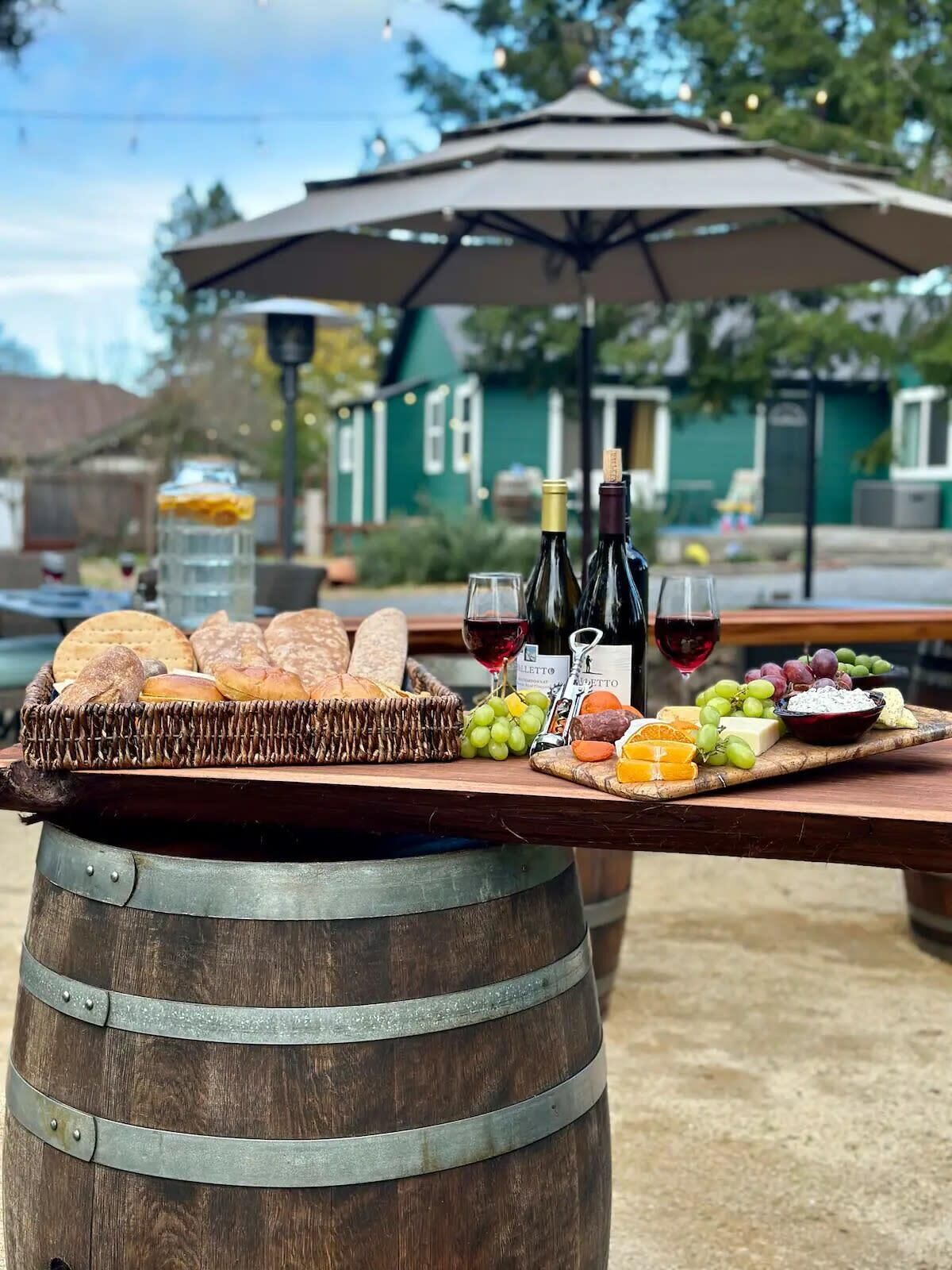 A wooden table with a wicker basket of bread, a cheese board with grapes, and two wine bottles.