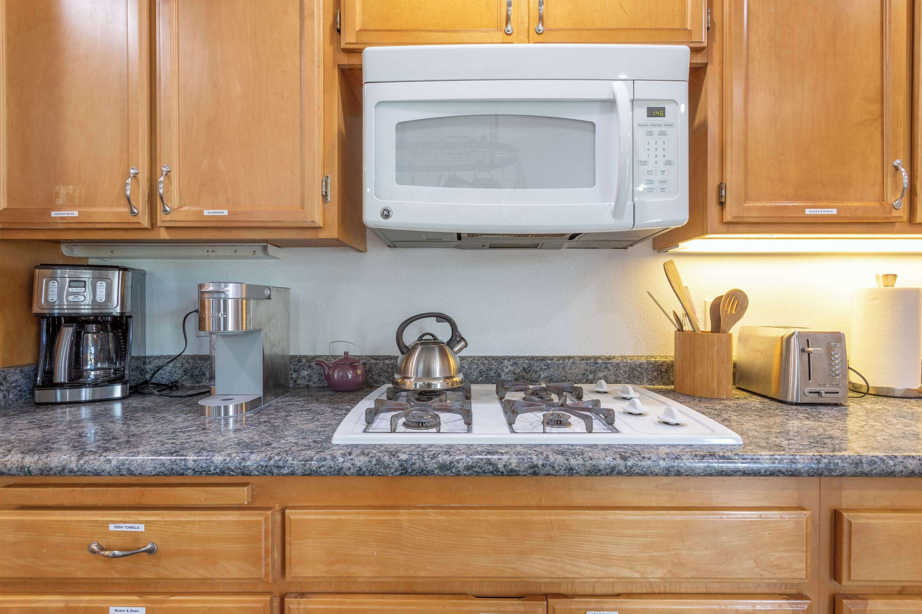 A kitchen with wooden cabinets, a white microwave, a gas stove, a coffee maker, a kettle, and a toaster.