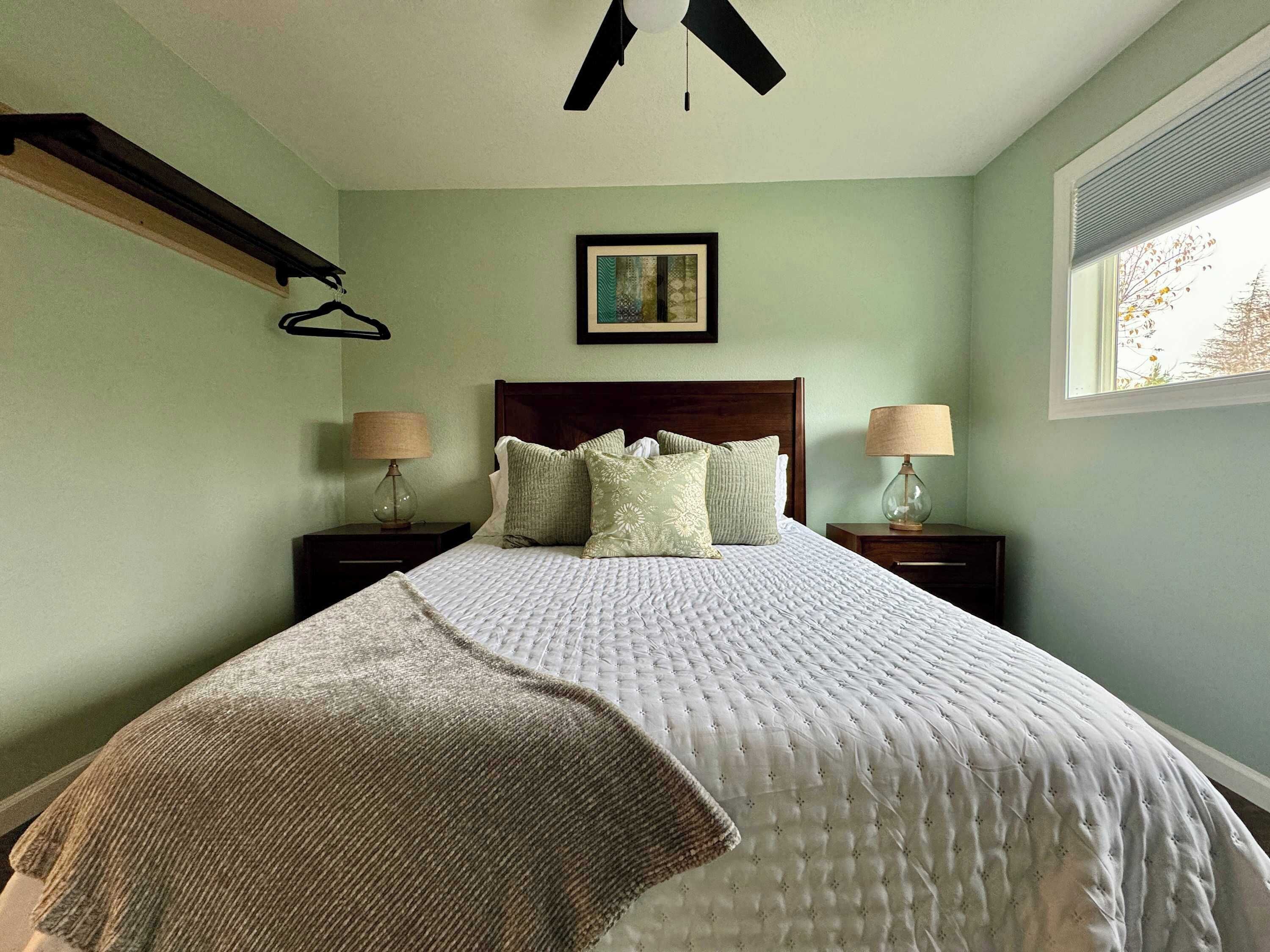 Una camera da letto con un letto, due comodini, una ventola a soffitto, una finestra e un quadro appeso al muro.