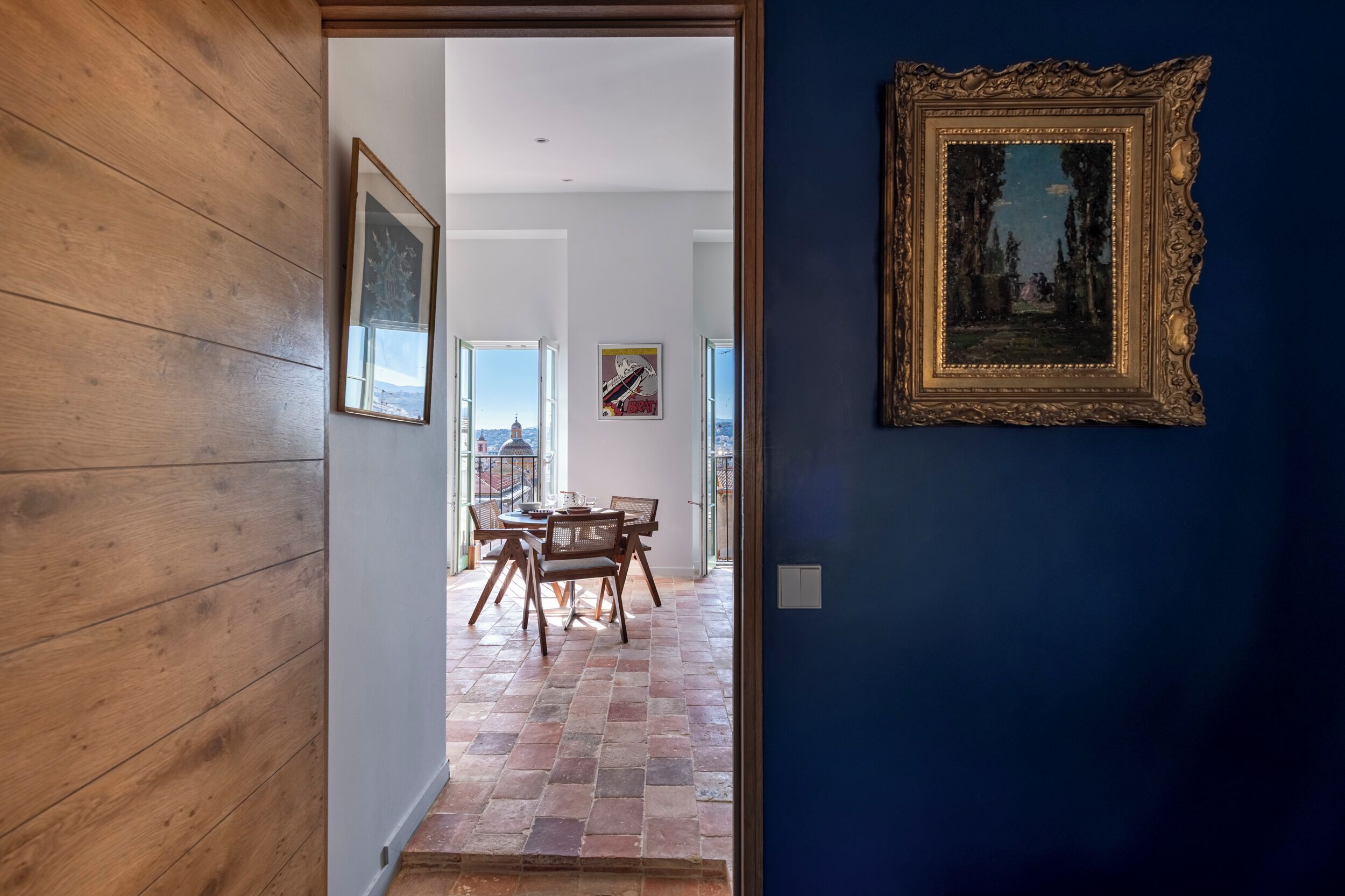 Une pièce avec un plancher en bois, une table à manger entourée de chaises, un tableau encadré au mur et une vue sur l’extérieur par une porte ouverte.