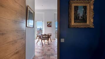 Une pièce avec un plancher en bois, une table à manger entourée de chaises, un tableau encadré au mur et une vue sur l’extérieur par une porte ouverte.