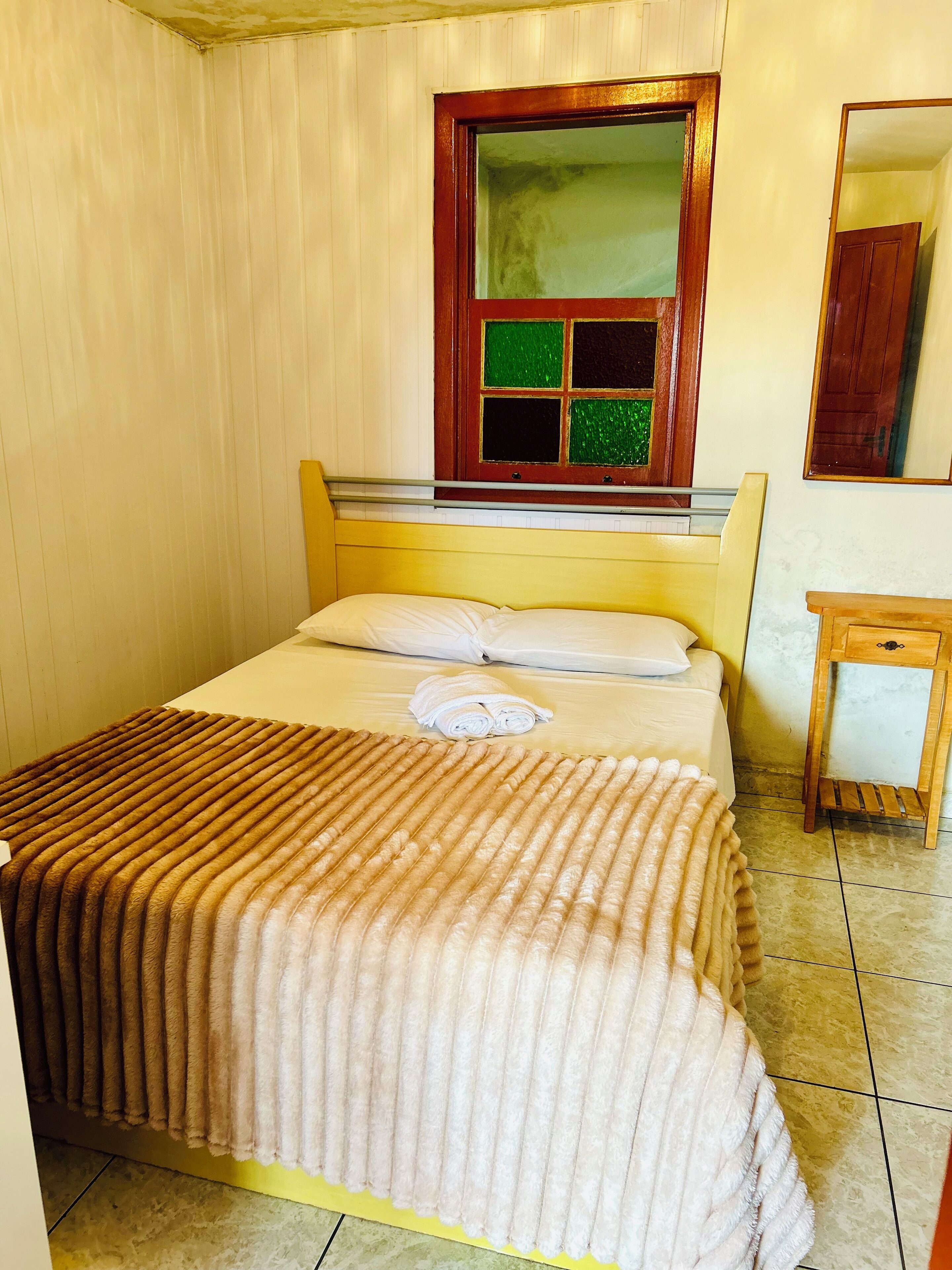 A single bed with a wooden headboard, a window with green and black panes, a small wooden nightstand, and a mirror on the wall.