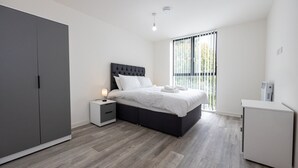 A modern bedroom with a large bed, a wardrobe, a bedside table with a lamp, and a view of greenery through the window. - GuestReady - Tranquil Vauxhall Stay (Liverpool)