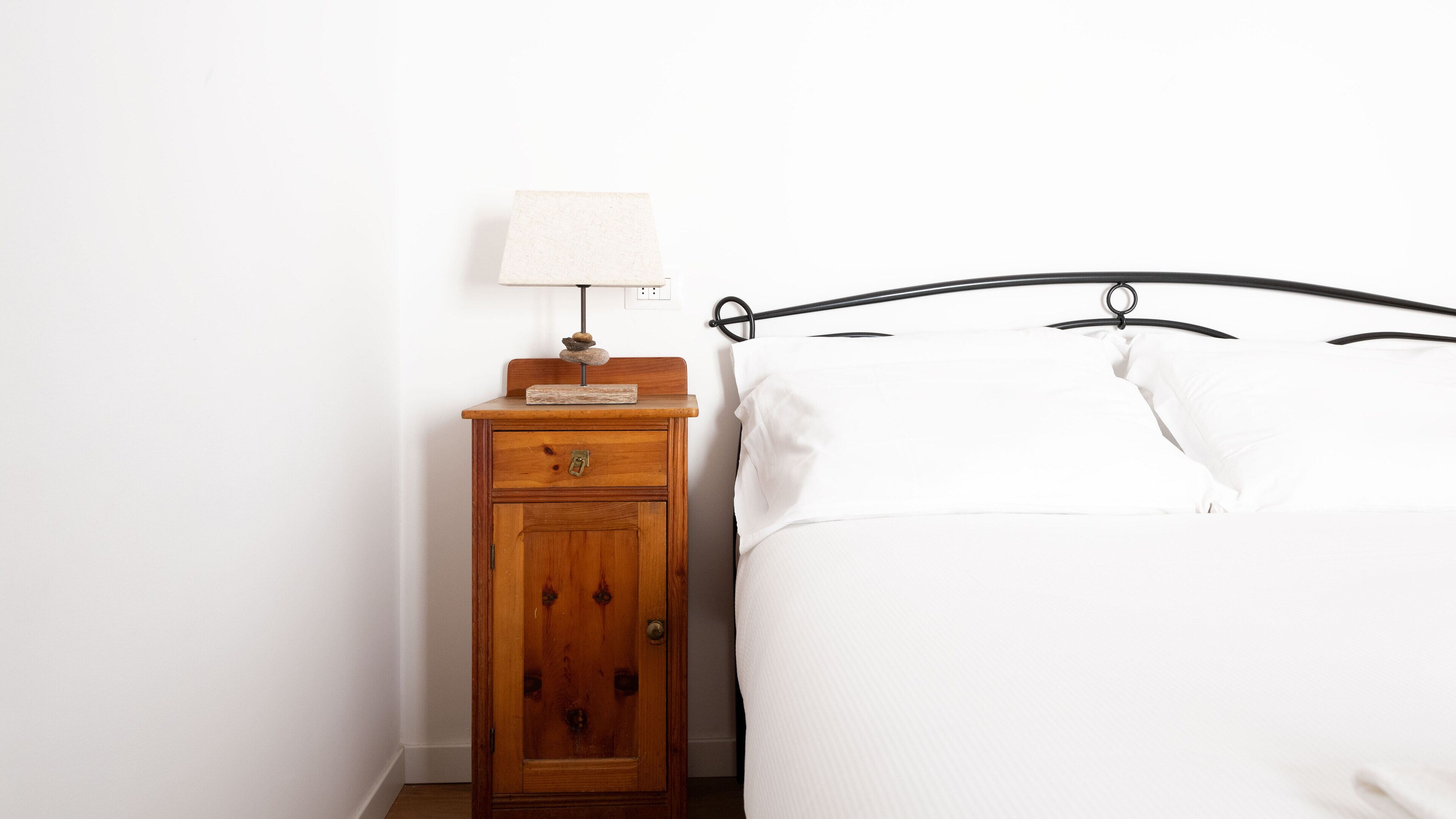 Ein Schlafzimmer mit Nachttisch aus Holz, einer Lampe und einem Bett mit weißer Bettwäsche.