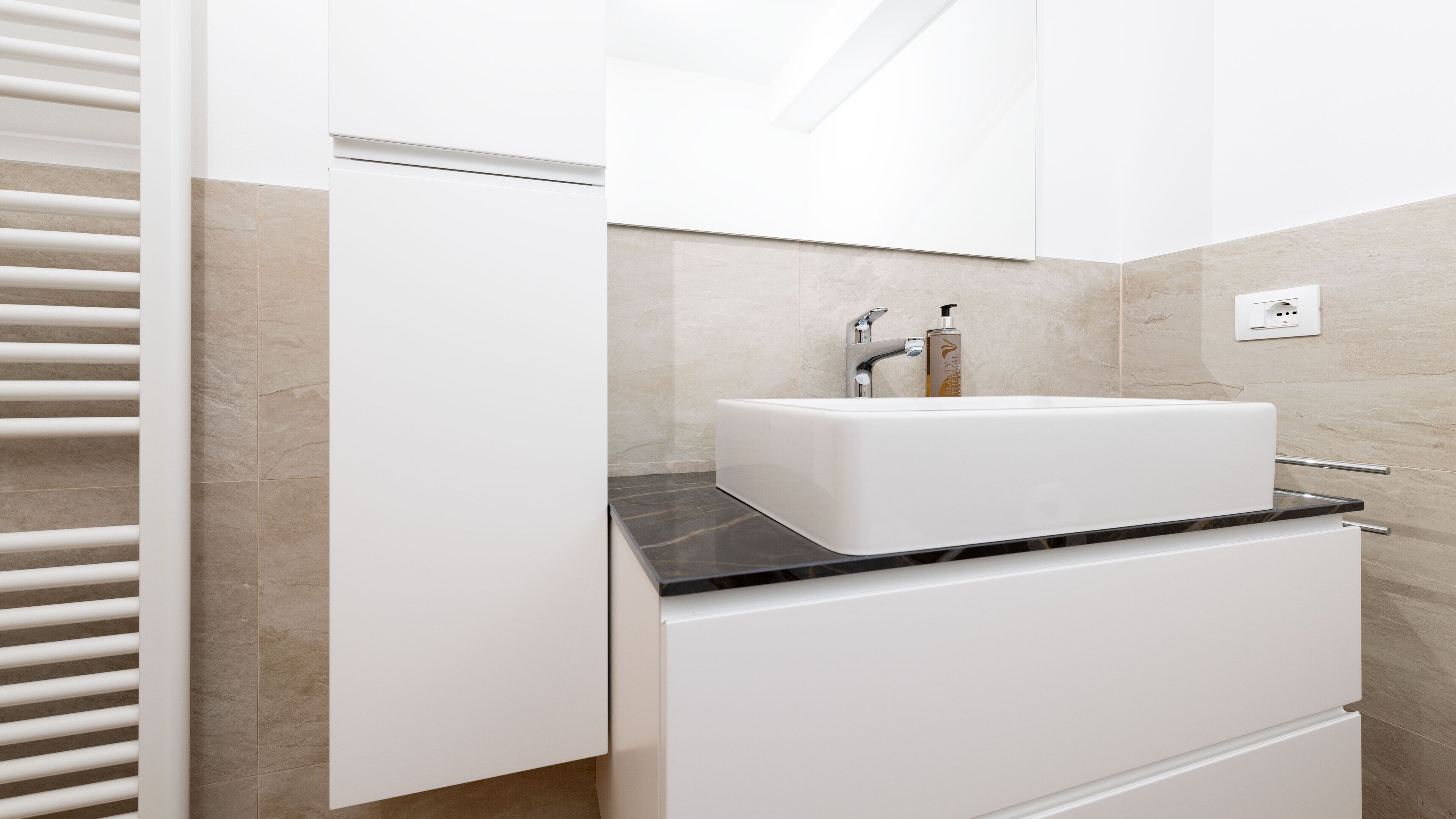 Un bagno moderno con un lavandino bianco, un piano di lavoro nero e pareti rivestite di piastrelle color beige.