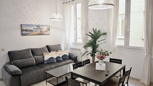 Una sala moderna con un sofá gris, una mesa de comedor con sillas, una planta en maceta y un cuadro en la pared.