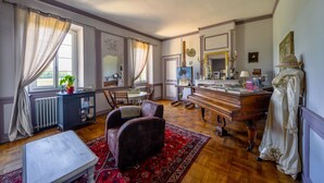 A spacious room with a wooden floor, a piano, a dining table with chairs, a large mirror, and a window with curtains.