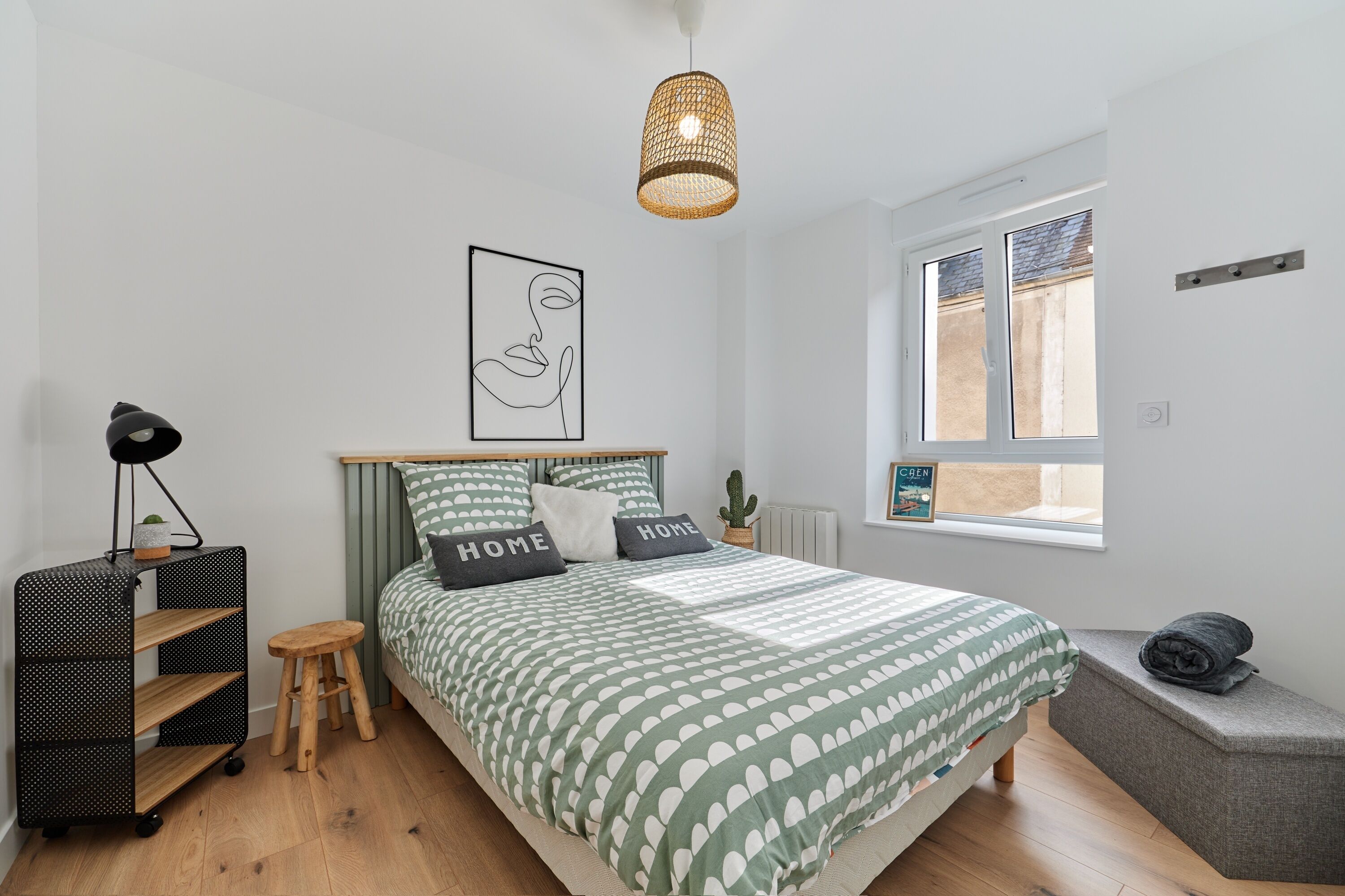 Ein Schlafzimmer mit Bett, Nachttisch, Hocker, Stehlampe, einer Regalwand, einem Fenster und einer Pendelleuchte.