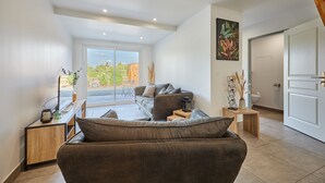 A modern living room with a sofa, a coffee table, and a TV stand. There is a door leading to another room and a window with a view of the outside.