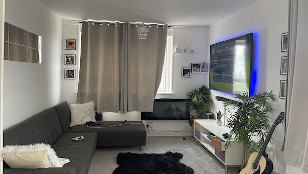 En moderne stue med en grå sofa, en sort, blød måtte, en guitar og et fjernsyn monteret på væggen.