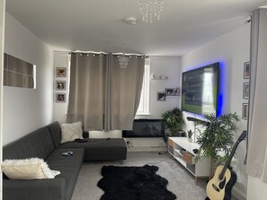 A modern living room with a grey sofa, a black furry rug, a guitar, and a TV mounted on the wall. - Stunning Welcoming Cosy Apartment in London (London)