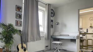 Et værelse med klaver, guitar, plante, et vindue med gardiner og et ur på væggen.