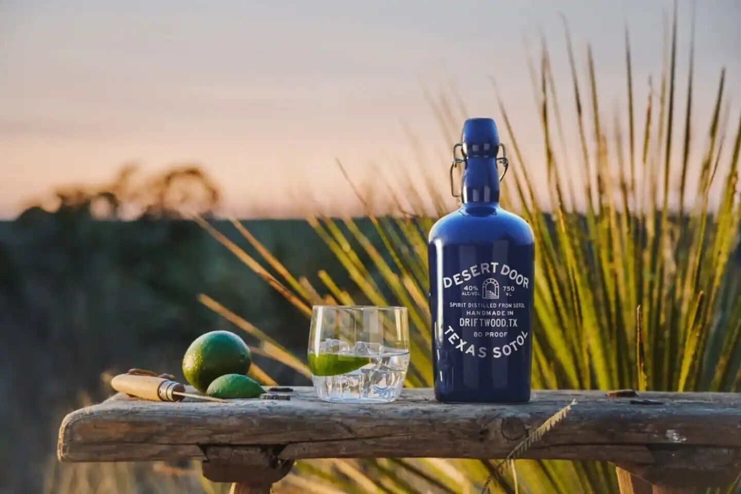 A bottle of Desert Door 750ml alcohol, a glass with a lime slice, and two limes on a wooden surface with a blurred natural background.