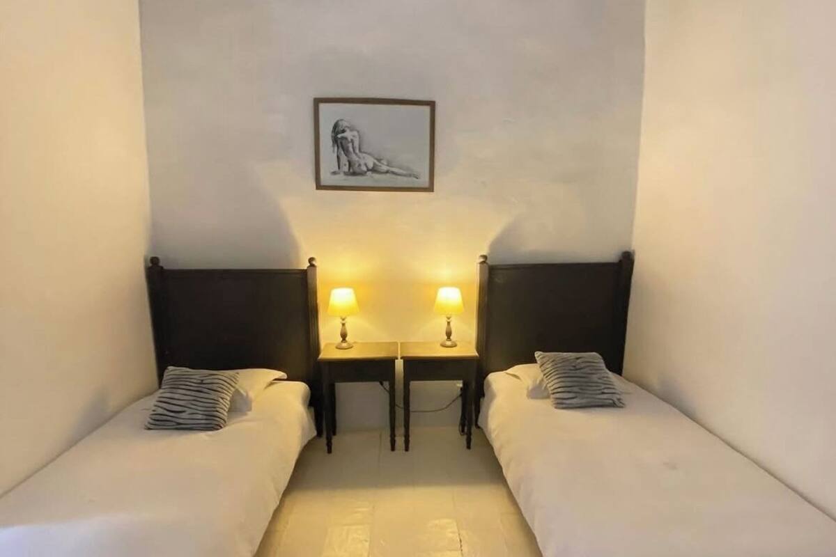 A room with two single beds, a small table, two bedside lamps, and a framed picture on the wall.