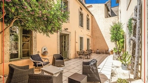 A courtyard with wicker furniture, a table, and chairs. There are potted plants and a tree with pink flowers. - Logement Touristique Villa Eugénie (Lezignan-Corbieres)
