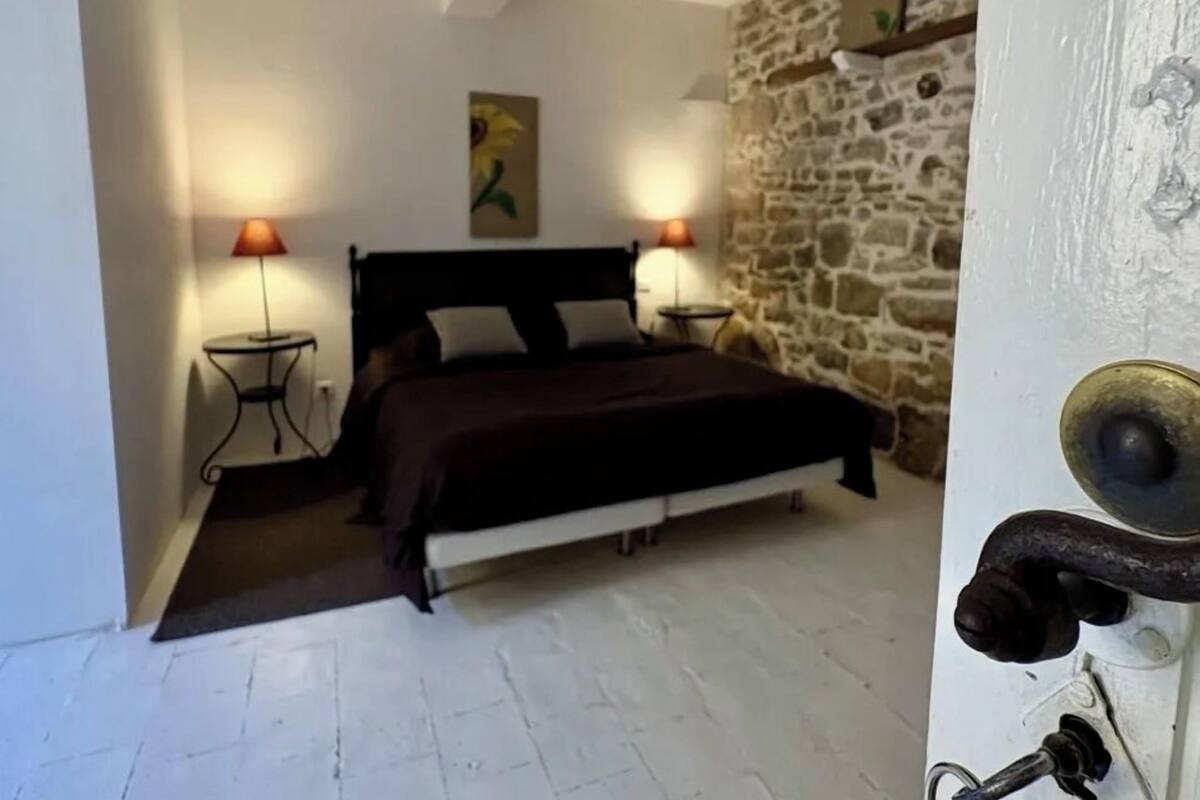 A bedroom with a bed, two bedside lamps, a painting, and a stone wall.