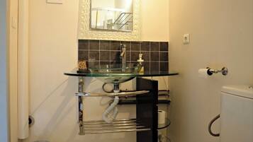 Une salle de bain avec un lavabo en verre, des toilettes, une poubelle et un porte-serviettes.