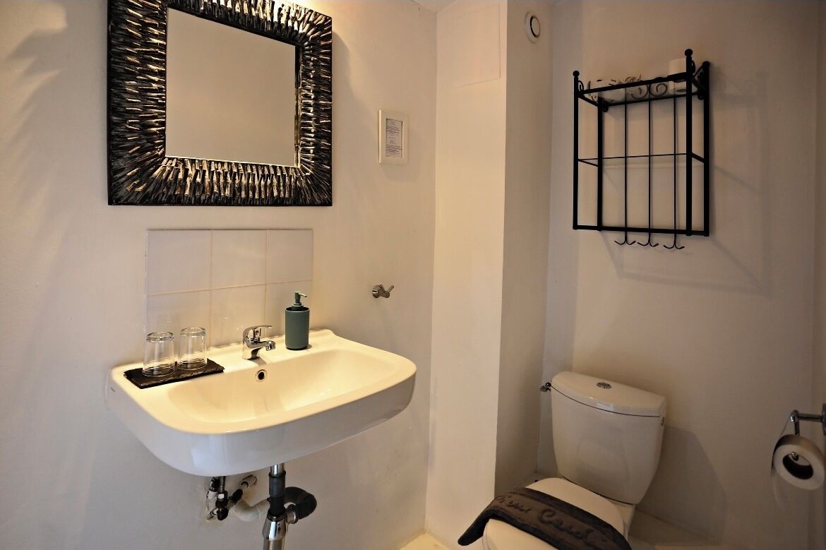 Un baño con un lavabo blanco, un espejo de marco texturizado, un inodoro y un estante de metal negro con un rollo de papel higiénico.