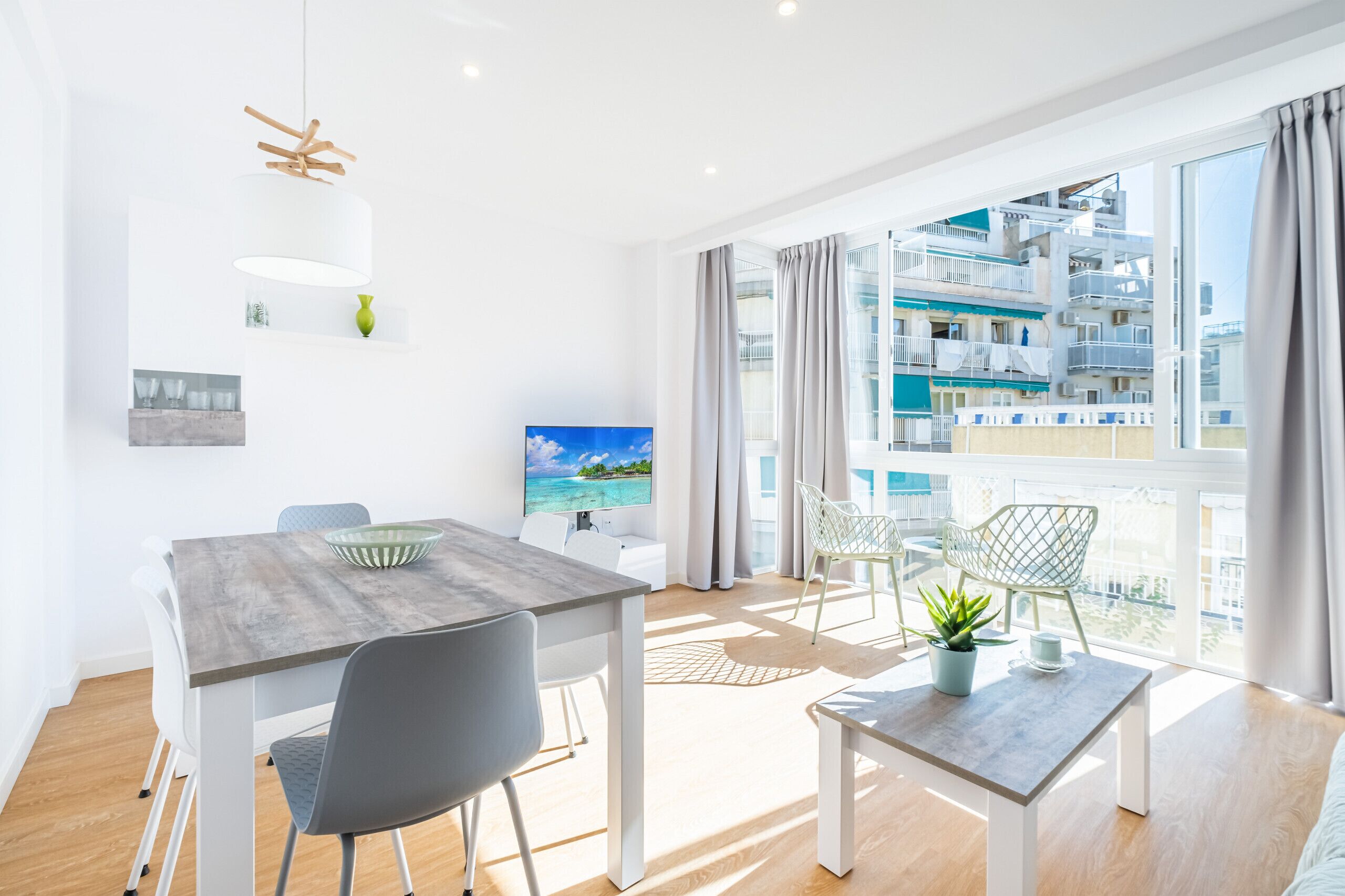 A modern living room with a dining area, large windows with a view of a building, and a flat-screen TV on the wall.
