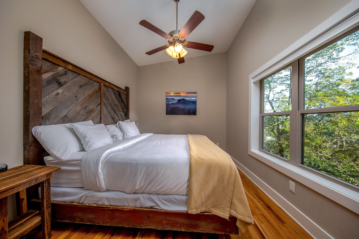 A bedroom with a wooden headboard, a bed with white linens, a ceiling fan, a window with a view of trees, and a small painting on the wall.