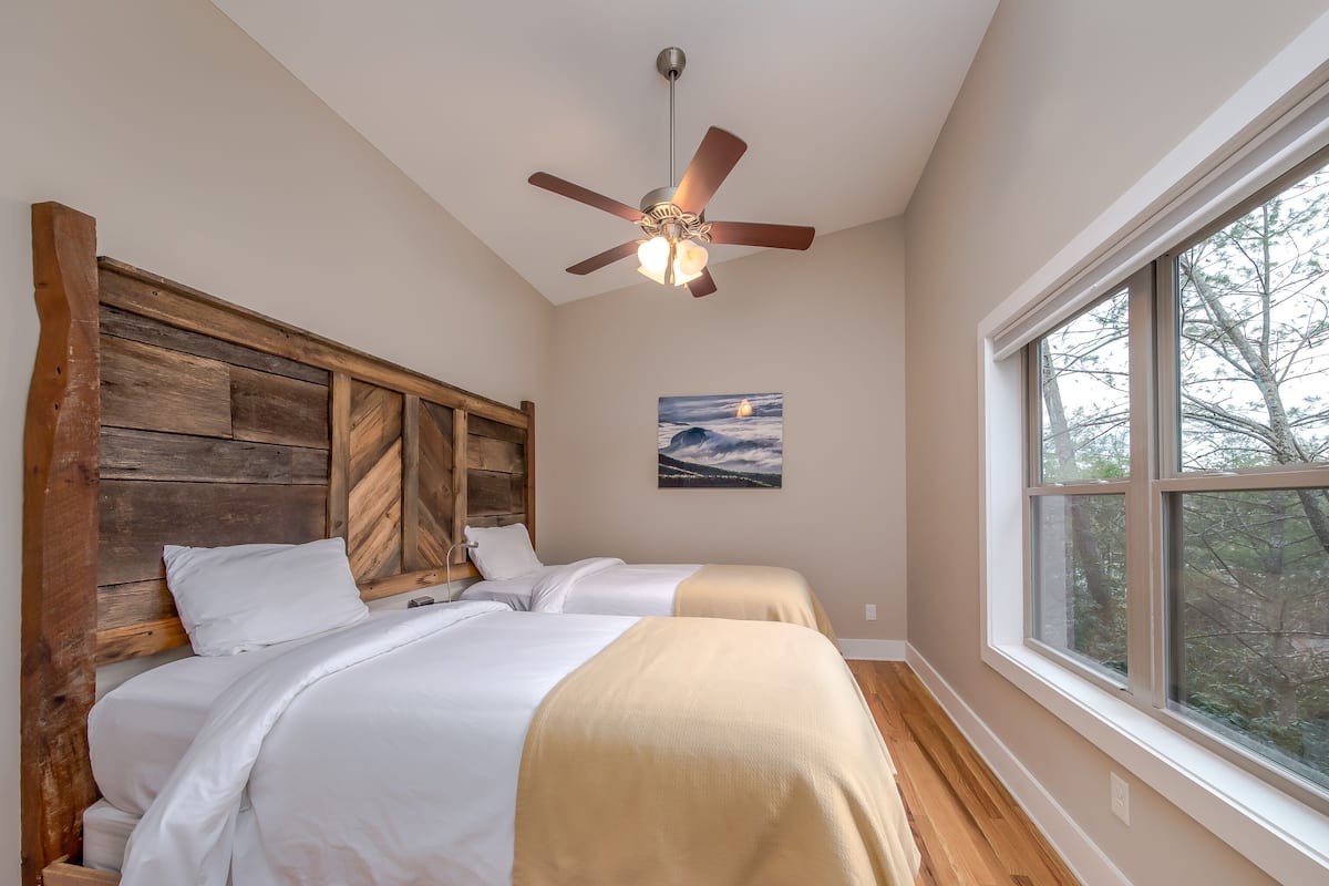 A bedroom with a wooden headboard, two beds, a ceiling fan, a painting, and a large window.