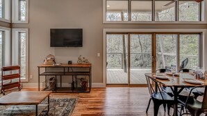 A spacious dining area with a large table set for six, a television mounted on the wall, and a view of the outdoors through large windows and sliding glass doors. - Pilot Cove Forest Lodging (Pisgah Forest)