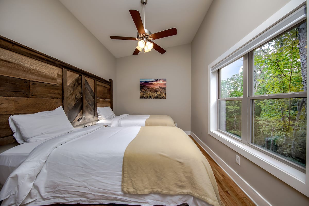 A bedroom with two beds, a ceiling fan, a large window with a view of trees, and a painting on the wall.