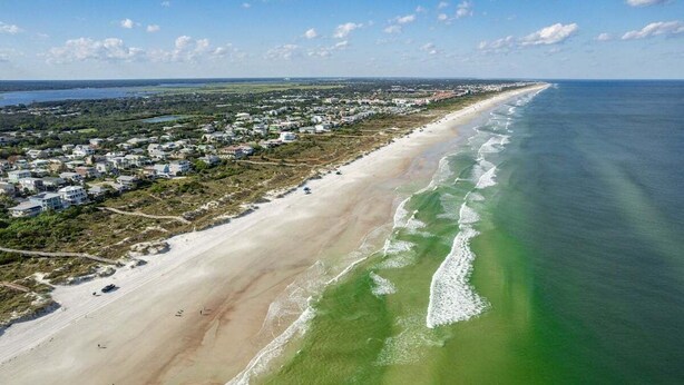 An aerial view of a coastal area with a beach, residential buildings, and a clear sky.