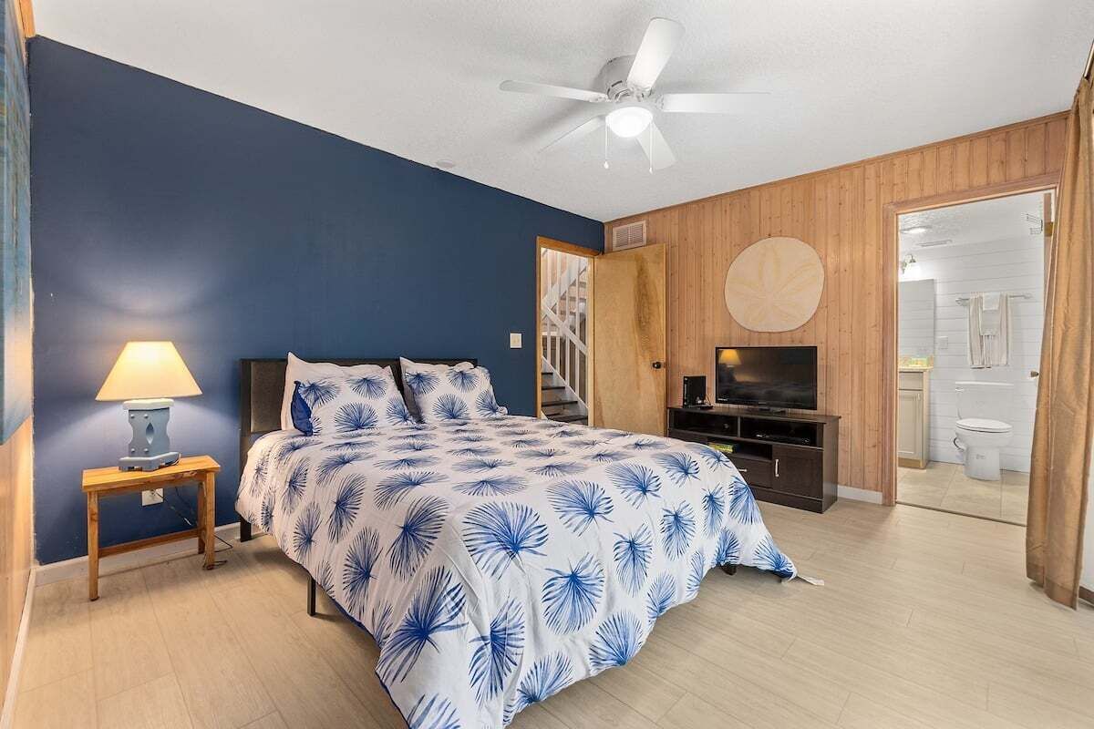 Quarto com cama, TV, ventilador de teto e porta de madeira que dá para um banheiro.