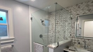 Ein modernes Badezimmer mit einer Duschkabine aus Glas, einem Waschtisch und einem Spiegel darüber. Ein Fenster mit einem Handtuch, das an der Tür hängt, und der Boden ist mit gemusterten Fliesen bedeckt.