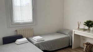 Un dormitorio con dos camas, un radiador, un equipo de aire acondicionado, un escritorio con cesta y una ventana con cortinas traslúcidas.