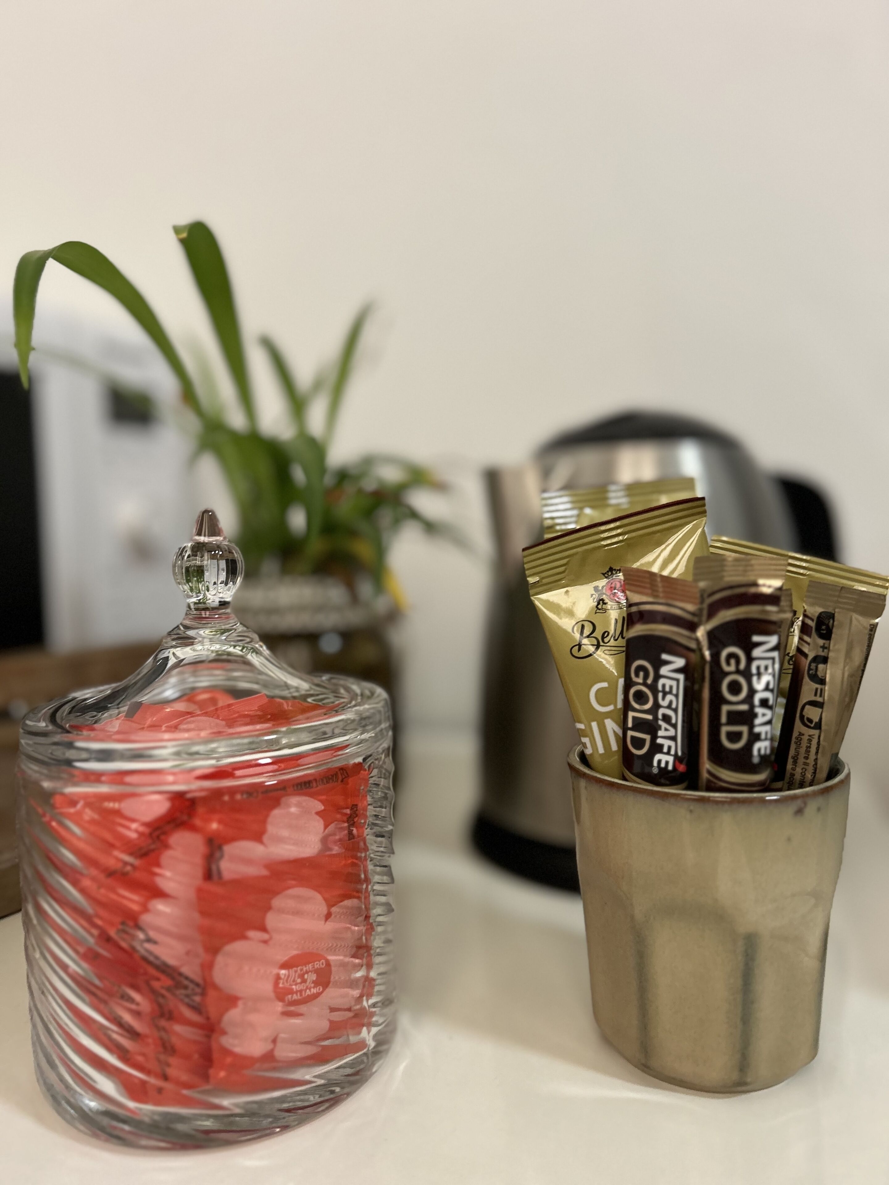 Een glazen pot gevuld met rode items, een metalen beker met Nescafe Gold pakjes, en een plantje op de achtergrond.