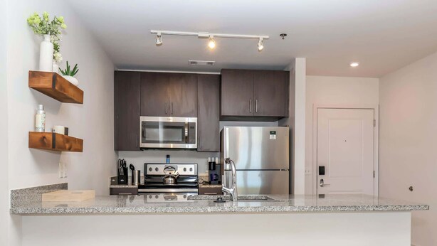 A modern kitchen with dark cabinets, stainless steel appliances, and a granite countertop. There are three wooden stools at the island.