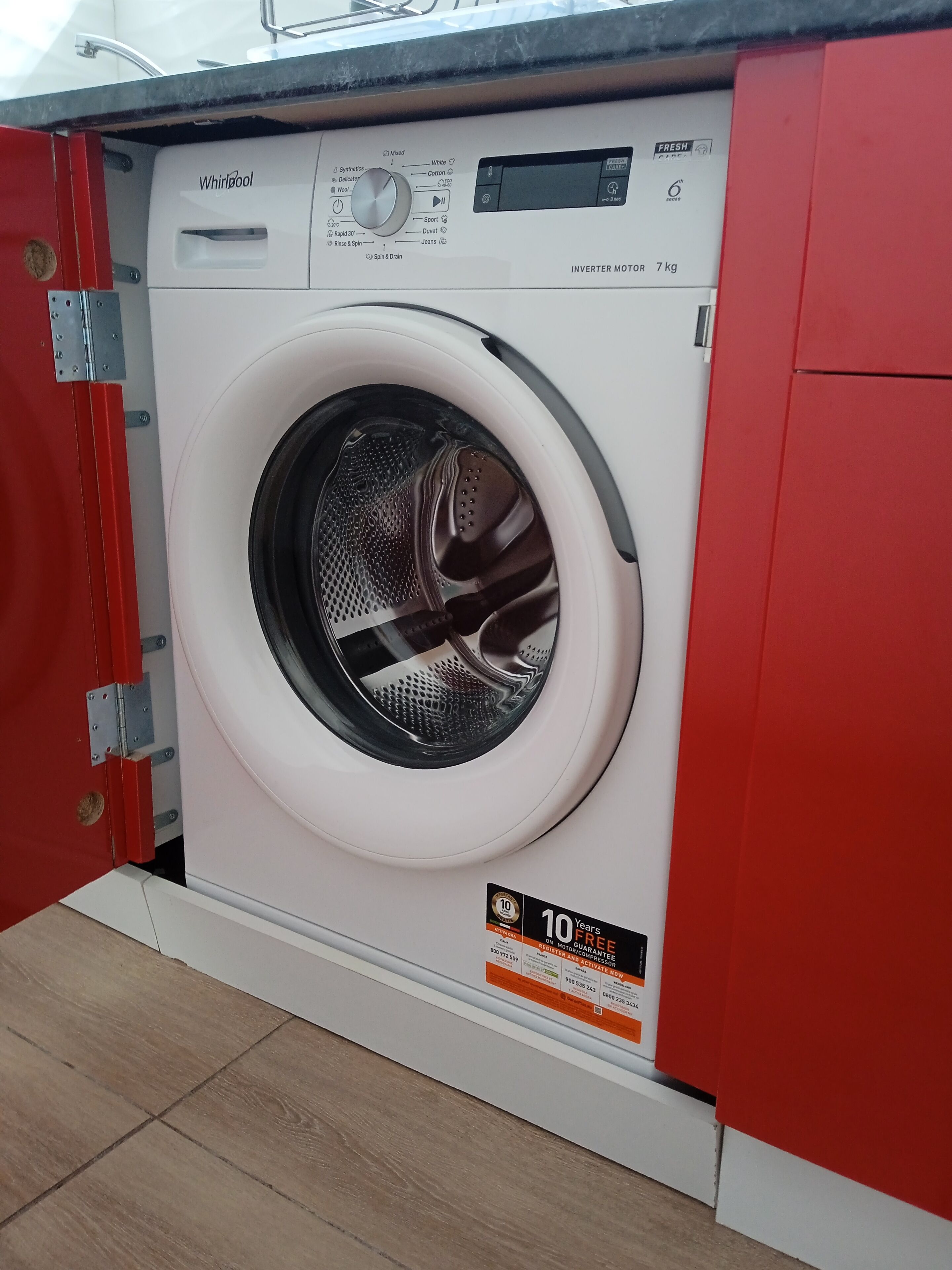 Una lavadora Whirlpool con un adhesivo de garantía de 10 años, ubicada en una cocina con un armario rojo.