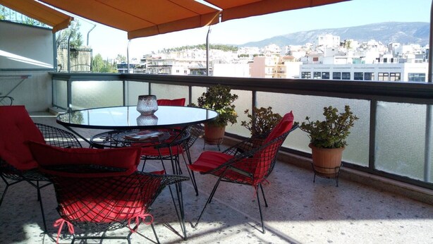 A balcony with a round table and chairs, potted plants, and a view of buildings and mountains. - Acropolis penthouse with breathtaking city view (Athina)
