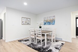 Uma sala de jantar com uma mesa de madeira e quatro cadeiras, um tapete preto e branco, e duas obras de arte emolduradas nas paredes.