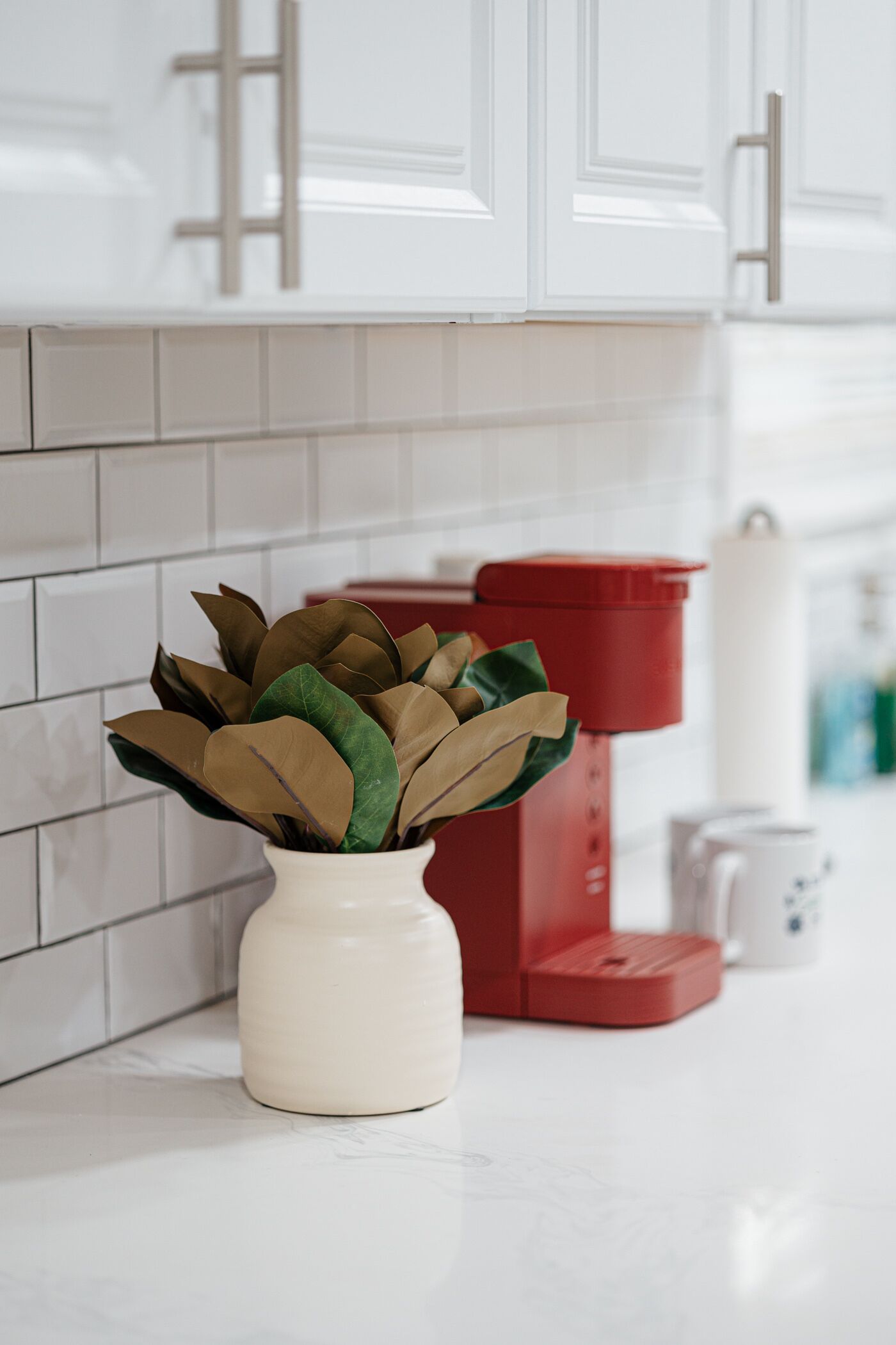 A kitchen with white cabinets, a white vase with artificial plants, and a red coffee maker on the counter.