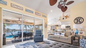 A spacious living area with a large sliding glass door leading to a pool, a comfortable sofa, a blue armchair, and a ceiling fan. - Luxury Tropical long waterfront view w/bikes,kayaks,golf clubs,beach gear incl. (Punta Gorda)