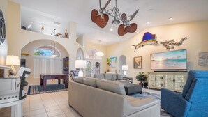 A spacious living room with a large sofa, a blue armchair, and a television mounted on the wall. There are decorative items on shelves and a ceiling fan with a unique design. - Luxury Tropical long waterfront view w/bikes,kayaks,golf clubs,beach gear incl. (Punta Gorda)