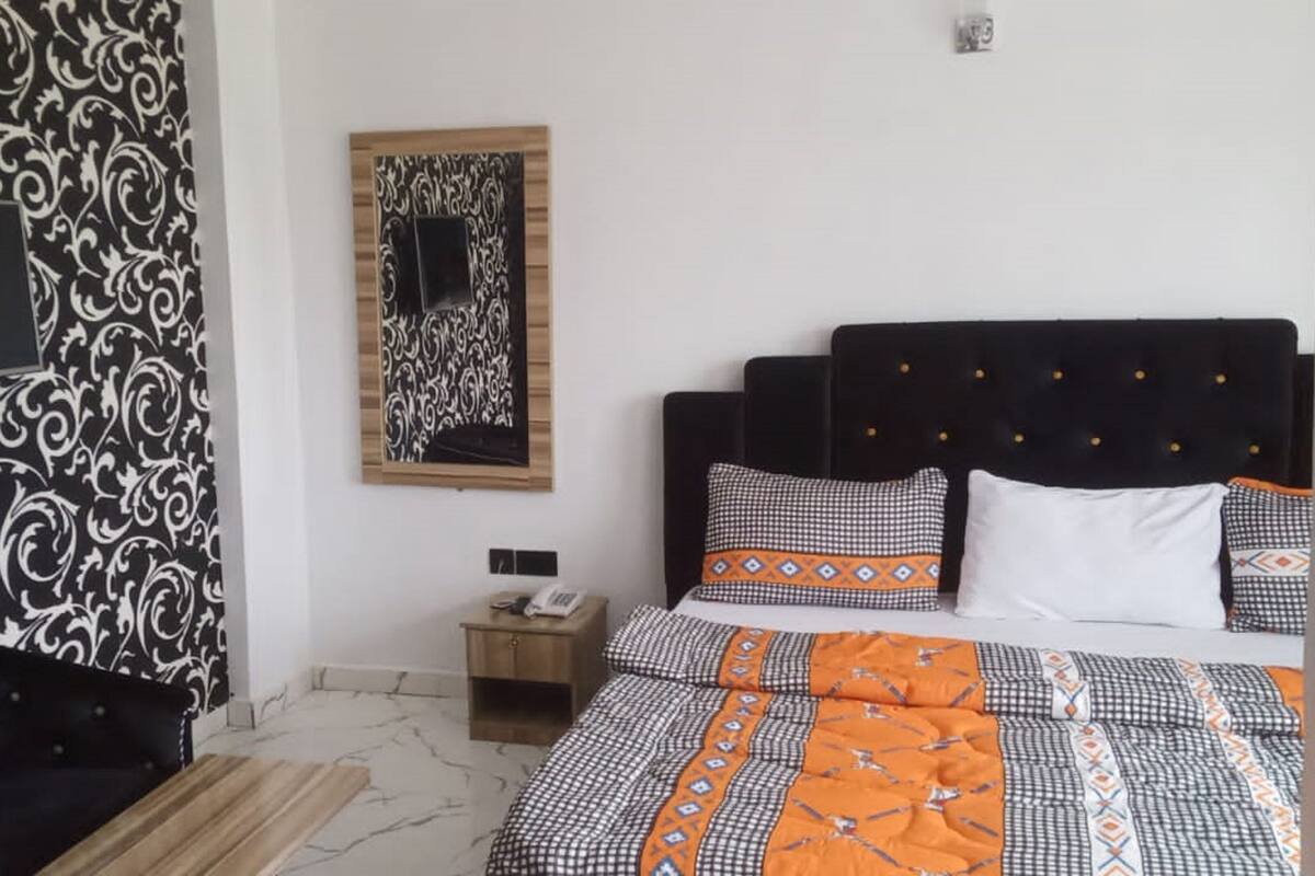 A bedroom with a patterned wall, a bed with checkered and orange bedding, a black upholstered headboard, and a small bedside table with a phone.