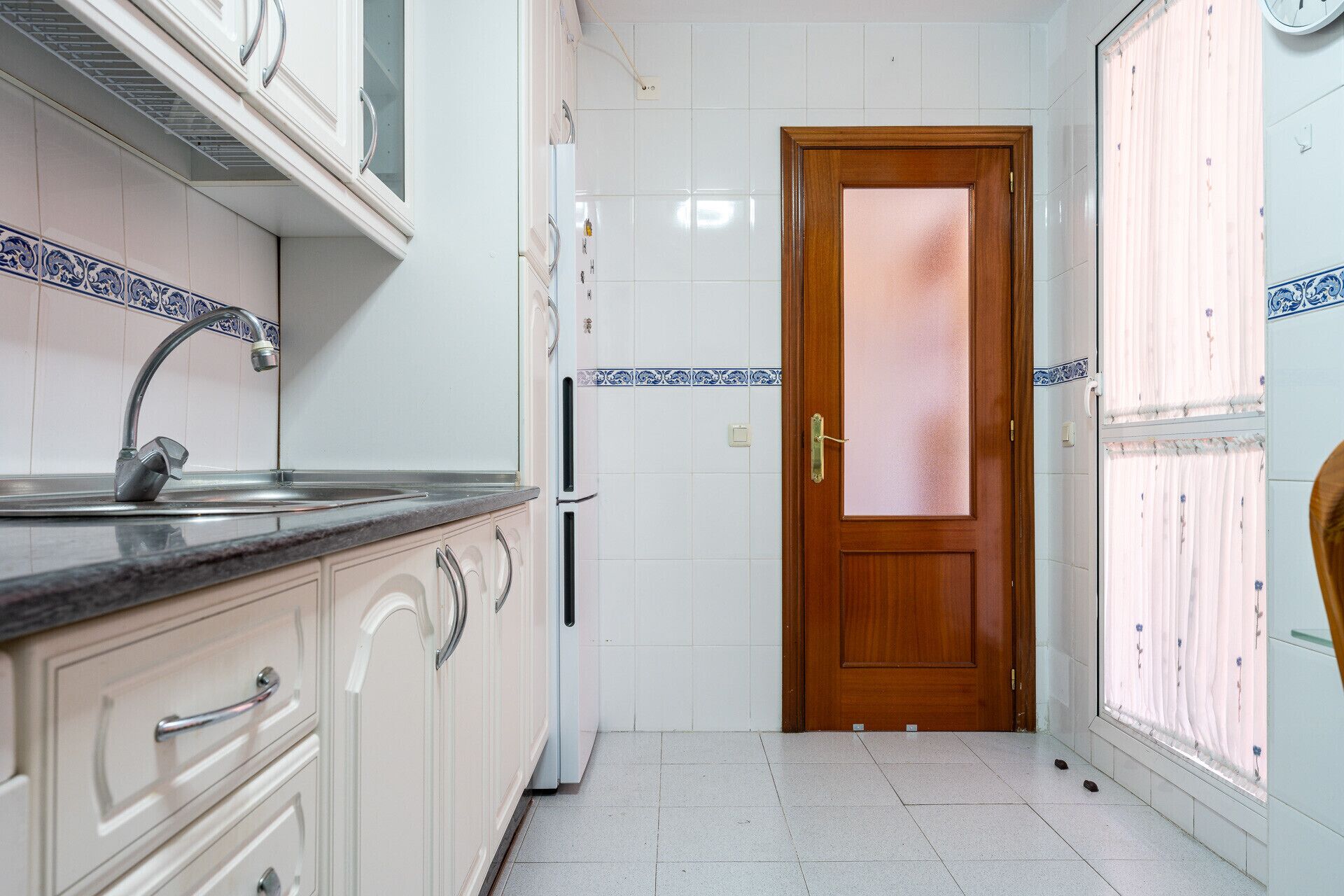 A narrow kitchen with white cabinets, a stainless steel sink, and a wooden door.