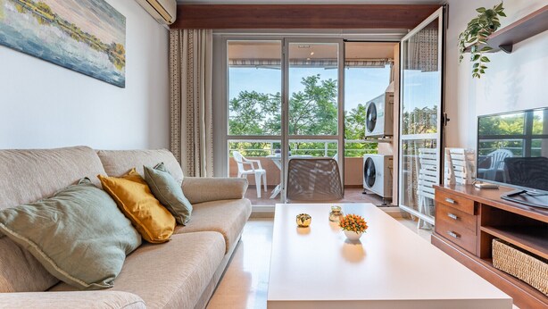Sala de estar con sofá, mesa de centro y mueble para televisión. Tiene un balcón con mobiliario exterior y vista a árboles.