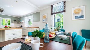 A modern kitchen with white cabinets, a dining table with teal chairs, and a large window with blinds. - Villa Moongate - Three Bedroom Villa, Sleeps 6 (Calahonda)