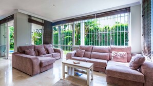A spacious living room with two sofas and a coffee table, large windows with metal bars, and a view of greenery outside. - Villa Moongate - Three Bedroom Villa, Sleeps 6 (Calahonda)