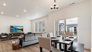 Una sala moderna con una televisión de pantalla plana, un sofá, una mesa de comedor con sillas y un amplio ventanal con vistas a casas vecinas.