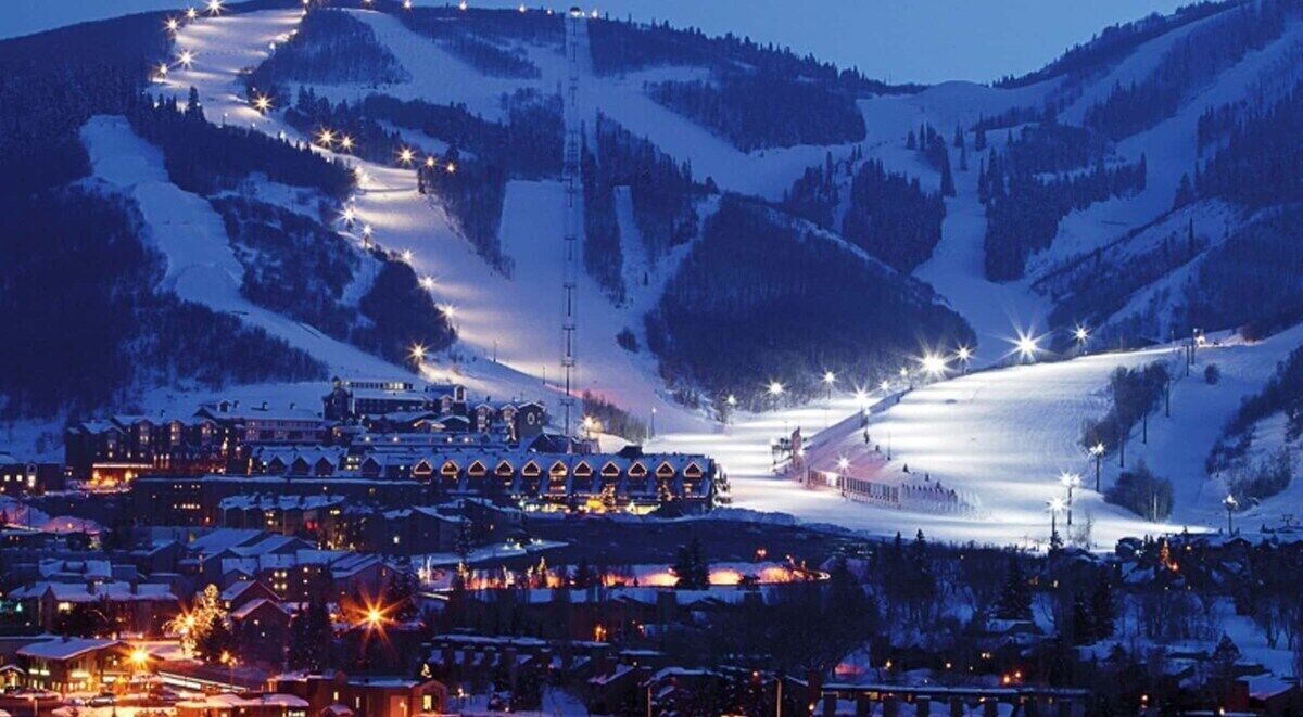 A snow-covered mountain resort at dusk with illuminated ski lifts, a central building with multiple windows, and surrounding snow-covered trees.