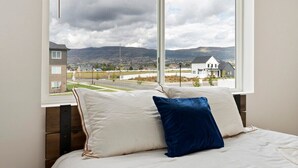 Un dormitorio con un ventanal grande, una cama con almohadas blancas y azules, y vistas a una zona residencial con casas y montañas.